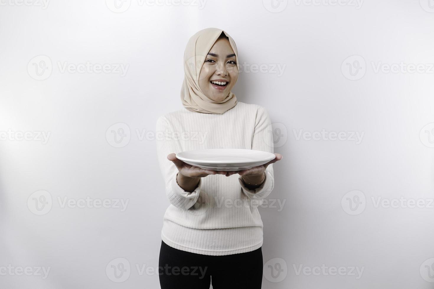 A smiling Asian Muslim woman is fasting and hungry and holding and pointing to a plate photo
