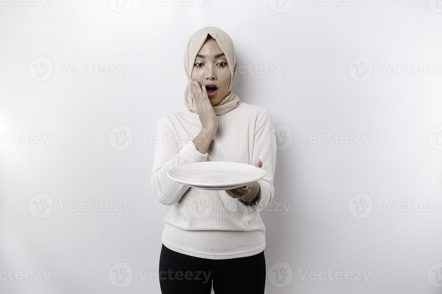 A shocked Asian Muslim woman is fasting and hungry and holding a plate with her mouth wide open photo