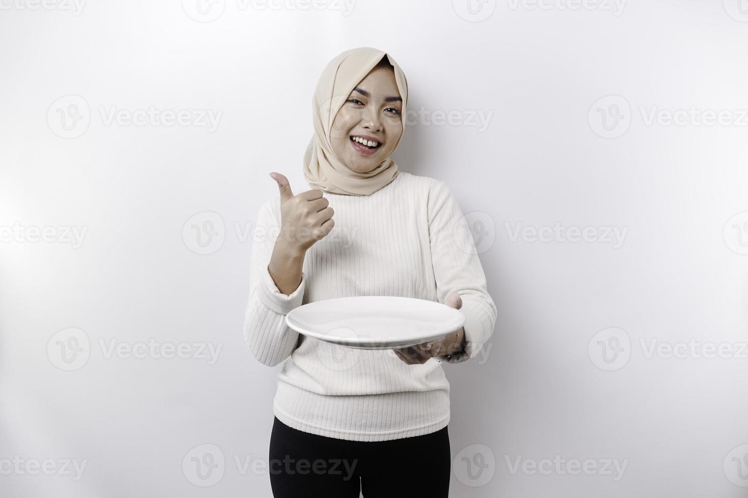 A smiling Asian Muslim woman is fasting and hungry and holding and pointing to a plate photo