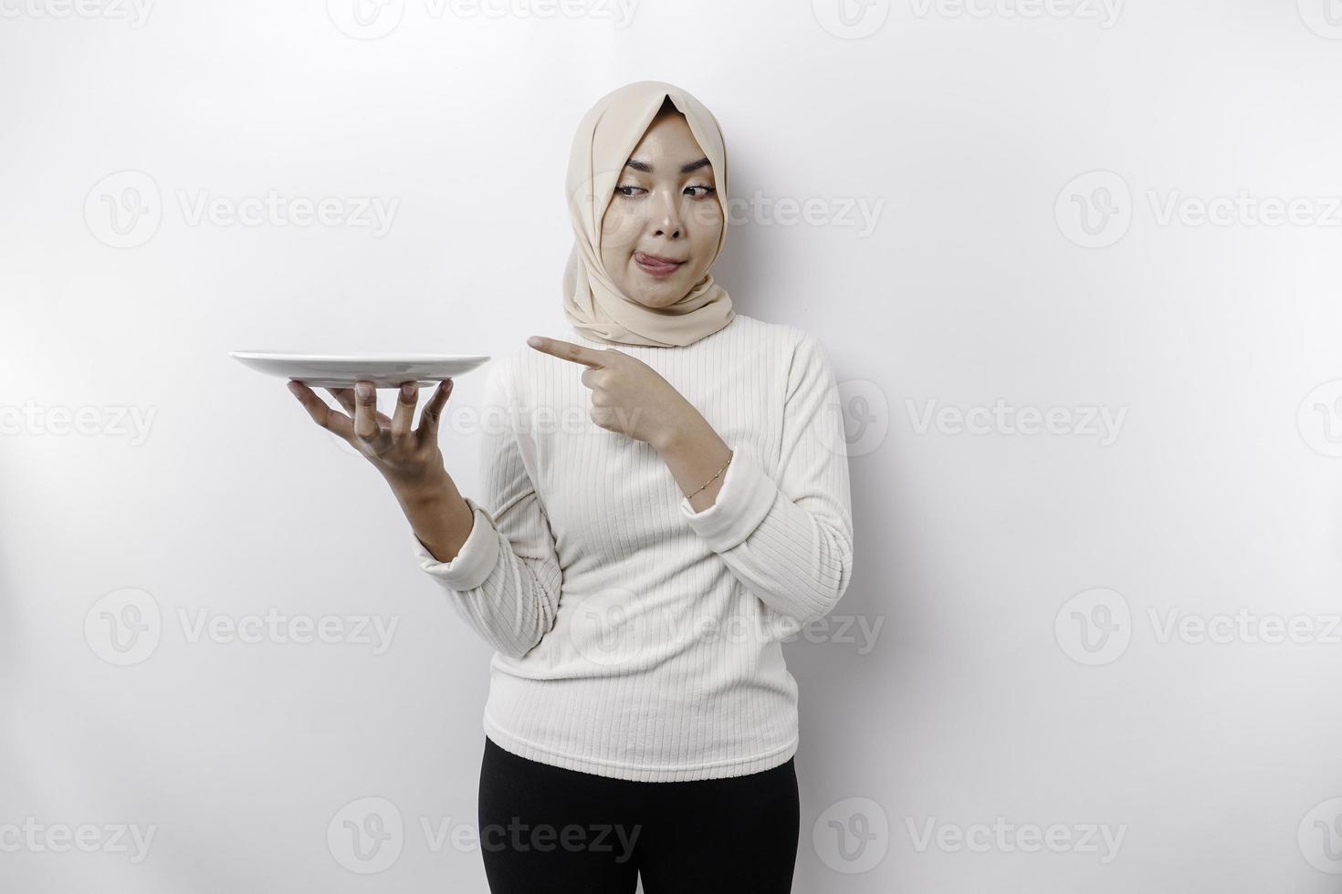 A smiling Asian Muslim woman is fasting and hungry and holding and pointing to a plate photo