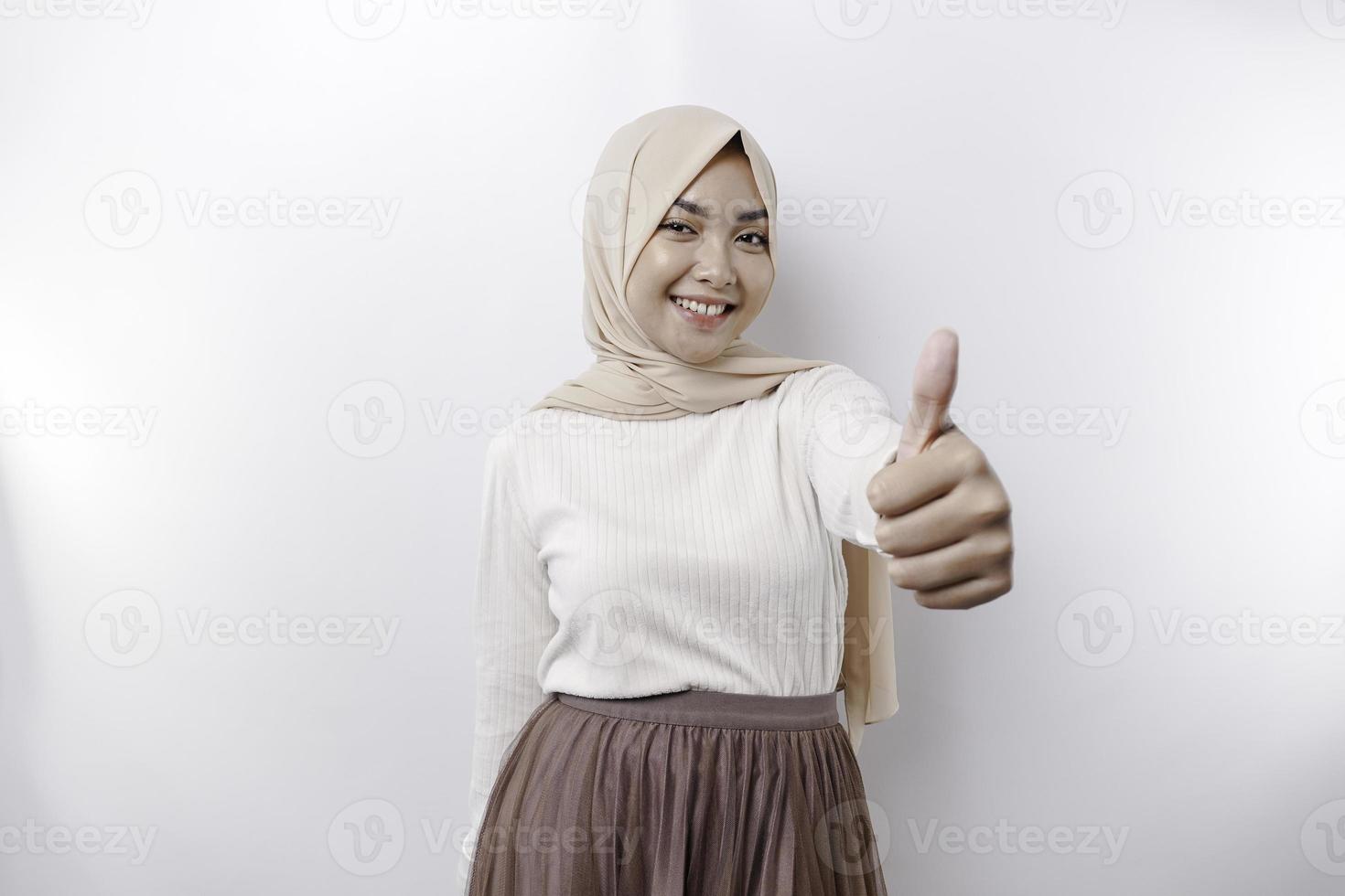 Excited Asian Muslim woman wearing a headscarf gives thumbs up hand gesture of approval, isolated by white background photo