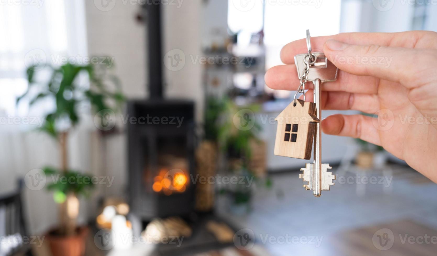 Key to house with keychain against background of fireplace stove with fire and firewood. Cozy home hearth. Building, design, project, moving to new house, mortgage, rent and purchase real estate photo