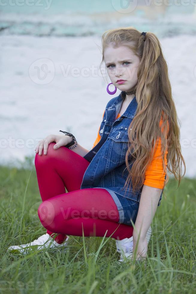 retrato de un pequeño Pelirrojo niña con brillante maquillaje en un mezclilla chaleco y rojo polainas. foto