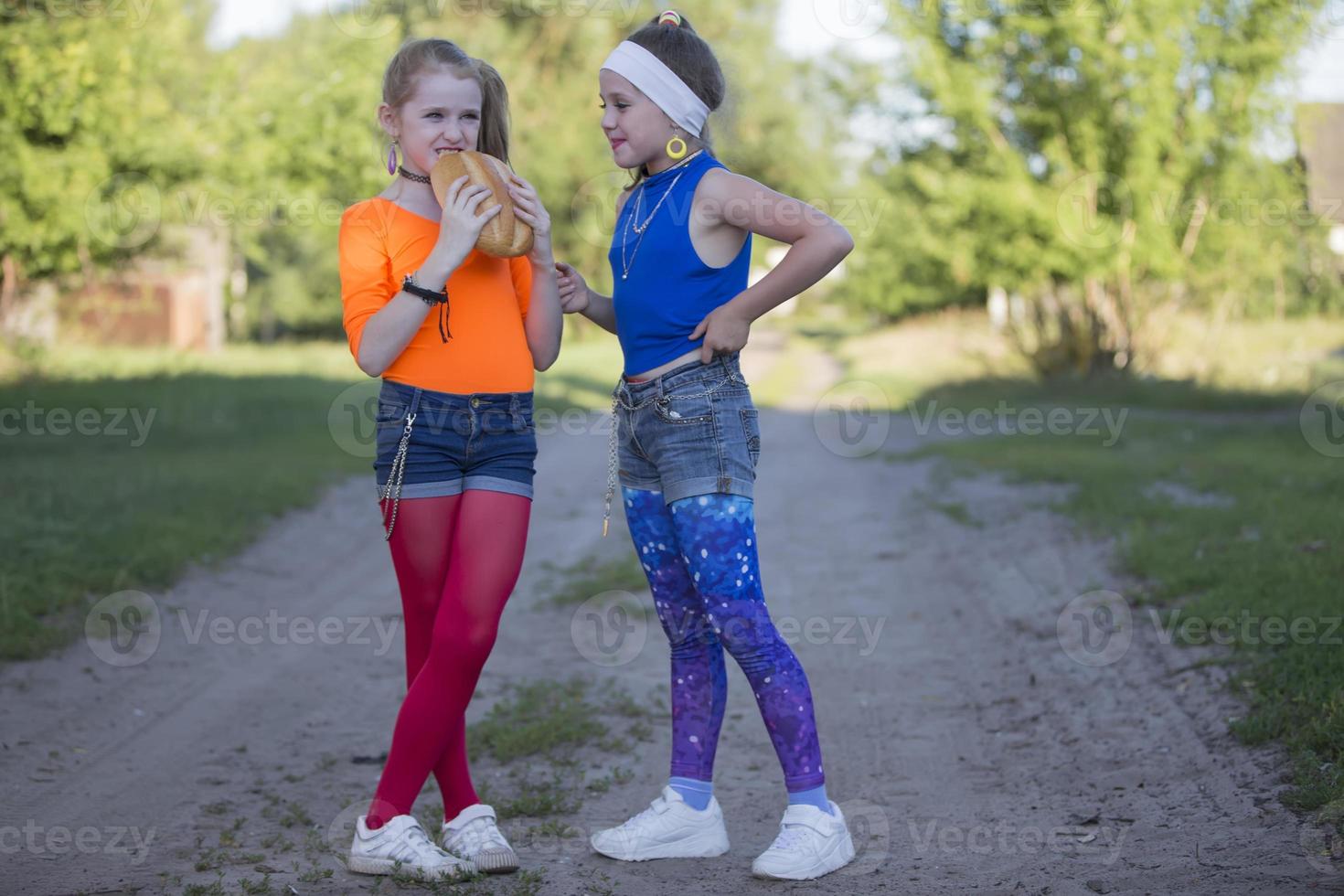Two girls with bright makeup dressed in the style of the nineties are eating a bun. Russian village children. photo