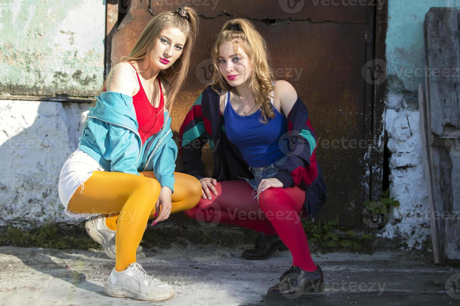 Two funny girls in bright clothes in the style of the nineties are sitting against the background of a rusty door. Russian village girls. photo