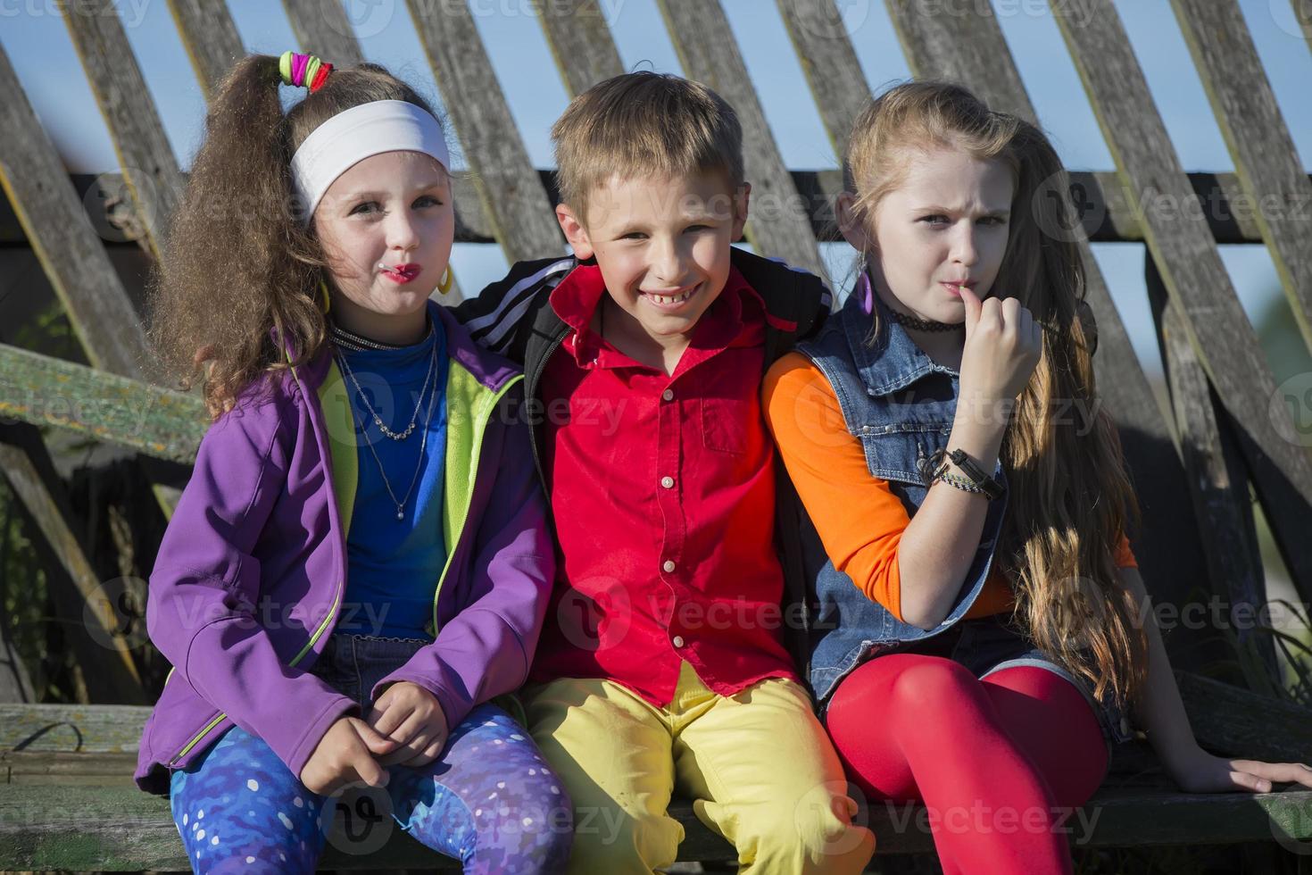 Funny little children girls with bright makeup dressed in the style of the nineties and a boy in a red shirt are sitting on a bench by a skewed fence. Russian village children. photo