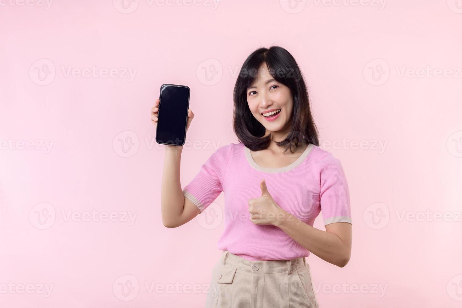 retrato hermosa joven asiático mujer contento sonrisa demostración teléfono inteligente pantalla con señalando dedo mano gesto a móvil teléfono aislado en rosado pastel estudio antecedentes. blanco pantalla teléfono inteligente concepto foto