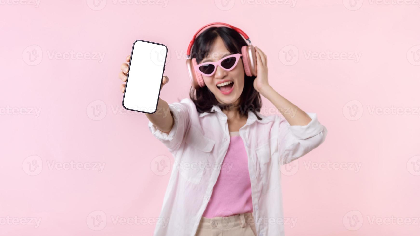 contento alegre sonriente asiático mujer con inalámbrico auriculares demostración blanco pantalla móvil teléfono o nuevo teléfono inteligente música solicitud anuncio Bosquejo aislado en rosado estudio antecedentes. foto
