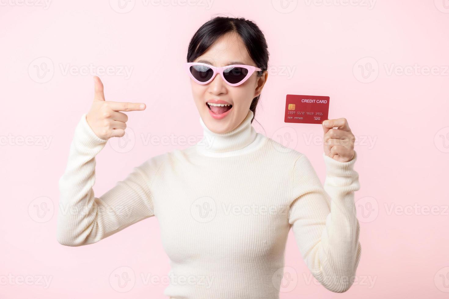 fascinante divertido alegre joven mujer de asiático etnia 20s años antiguo con vestir Gafas de sol usa blanco camisa sostener en mano crédito banco tarjeta aislado en llanura pastel ligero rosado antecedentes estudio retrato. foto