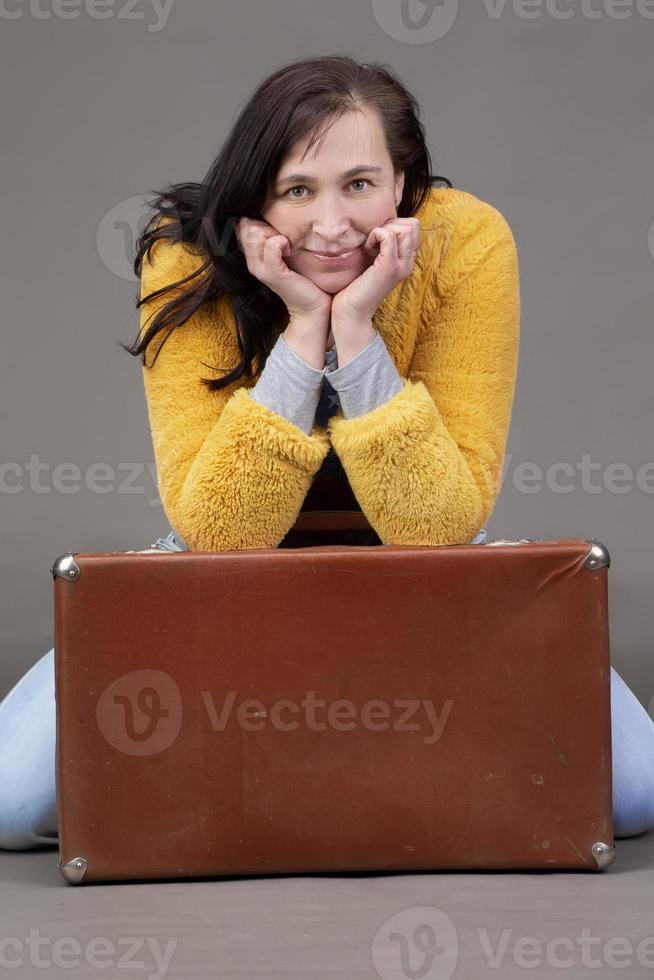 Happy fifty year old woman in yellow clothes with vintage suitcase on gray background. Portrait of a middle-aged woman. photo