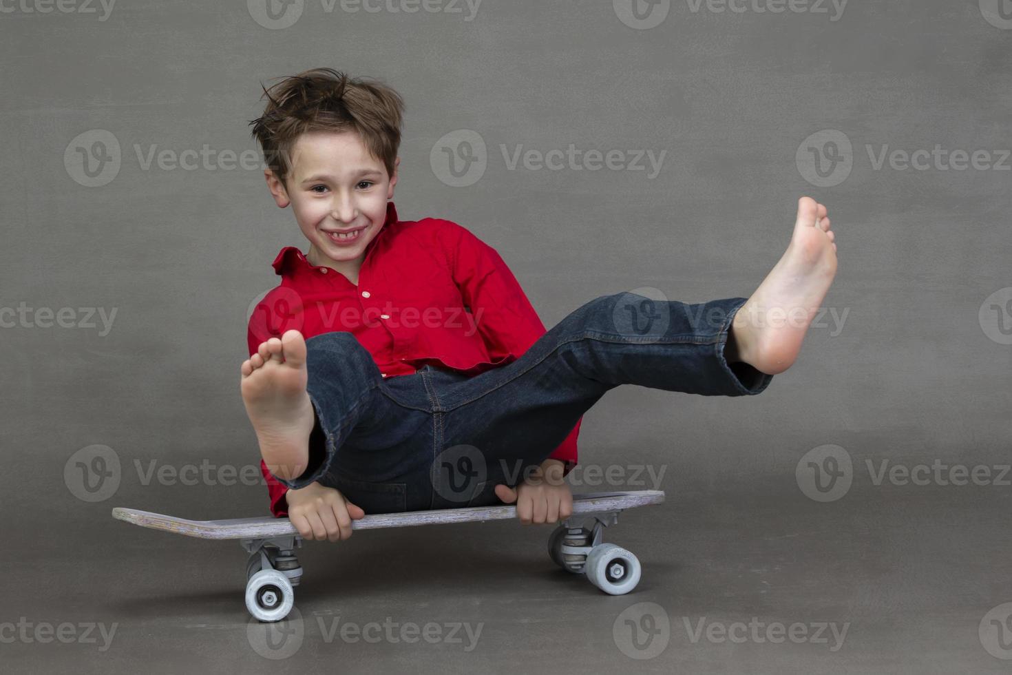 Funny little boy sitting on a vintage skateboard barefoot on a gray background. photo