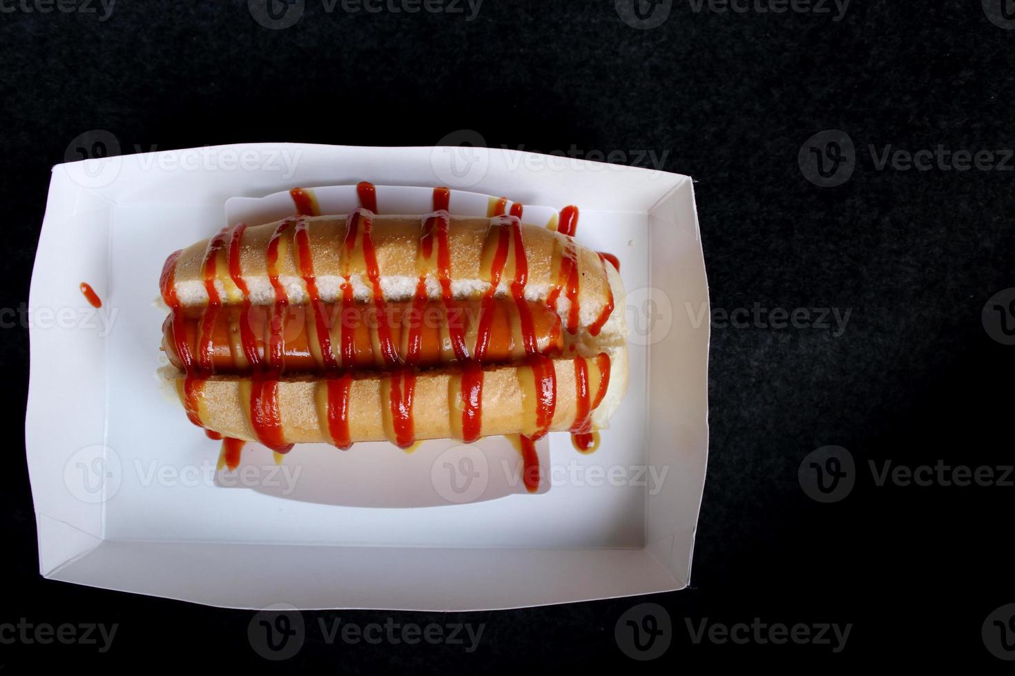 Hot dog in the slit of a partially sliced bun and top with tomato sauce, mayonnaise put in white paper box for take home and black background. photo