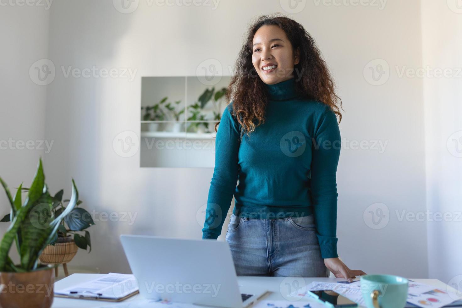 Smiling young Asian female employee stand at desk in office look in distance thinking or visualizing career success. Happy businesswoman plan or dream at workplace. Business vision concept. photo