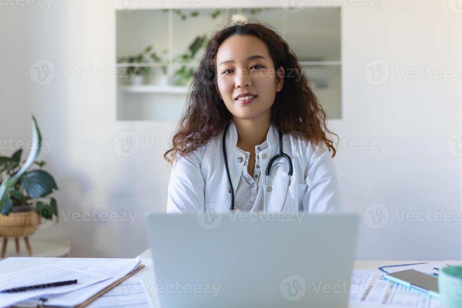youn asiático médico utilizando ordenador portátil y sentado a escritorio. mujer profesional médico médico vistiendo blanco Saco y estetoscopio trabajando en computadora a lugar de trabajo. foto