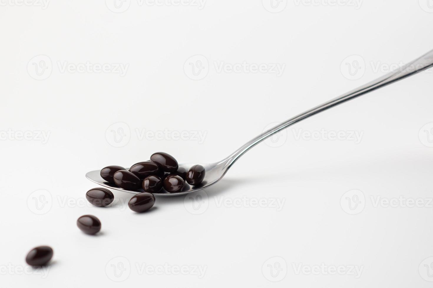 Black soft gel capsules in a stainless steel spoon on a white background. Vitamin food supplements photo