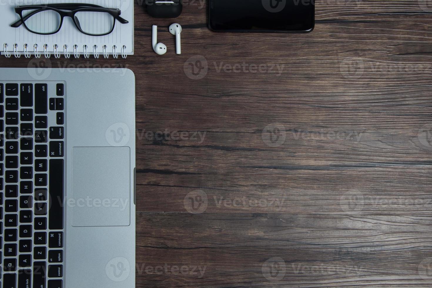wooden office desk with laptop, blank notebook, cordless earphones and glasses, dark background, wooden background, Top view with copy space, empty space. photo