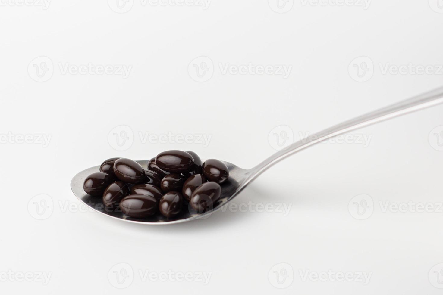 Black soft gel capsules in a stainless steel spoon on a white background. Vitamin food supplements photo