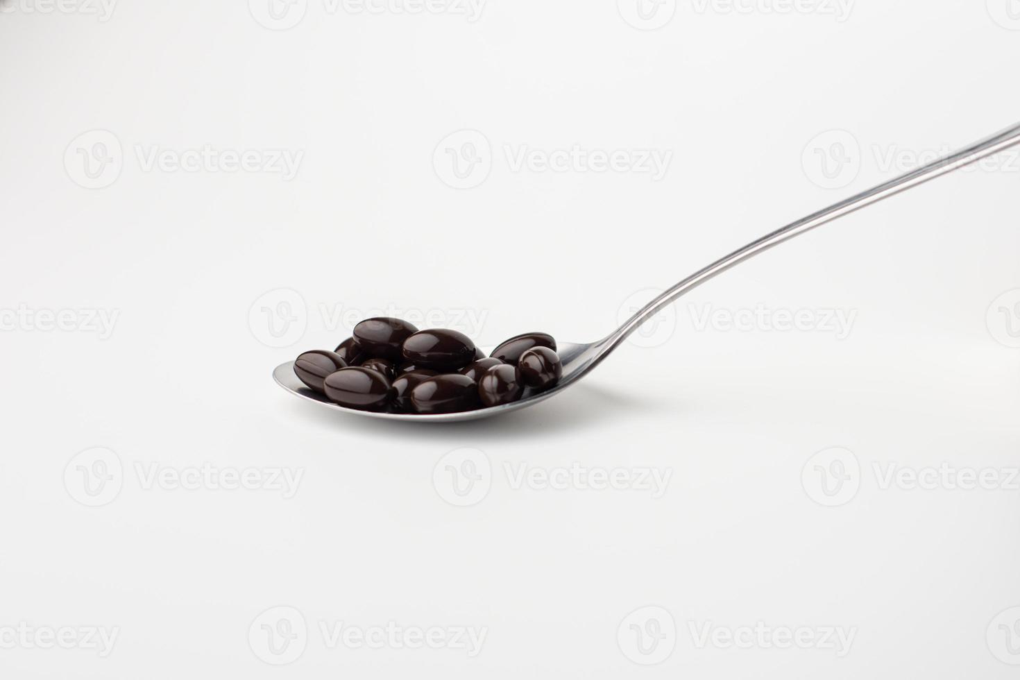 Black soft gel capsules in a stainless steel spoon on a white background. Vitamin food supplements photo