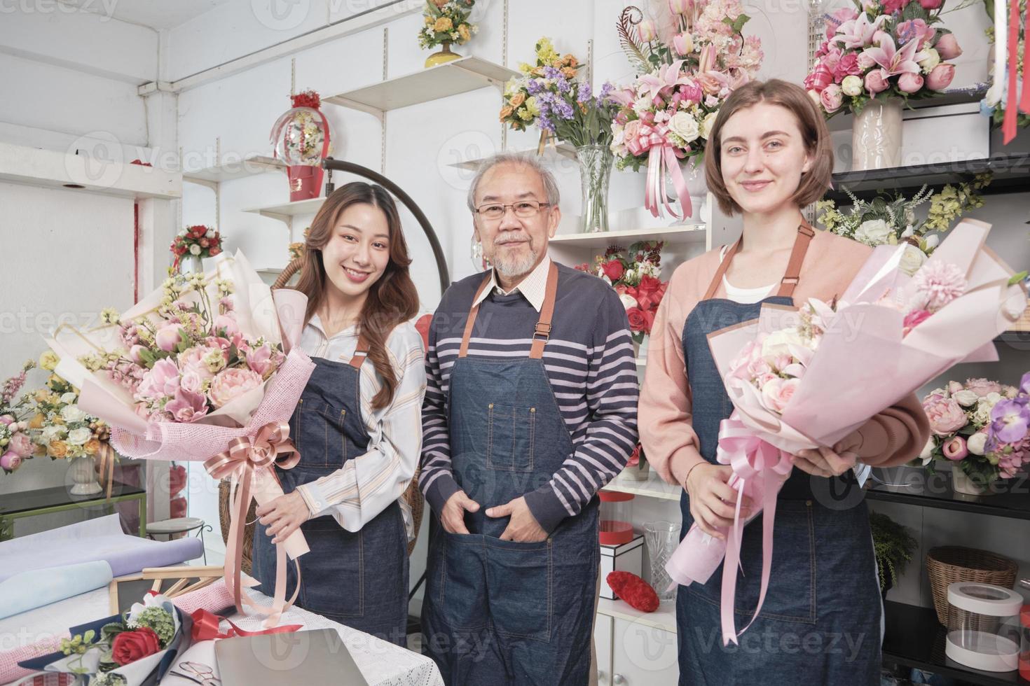 retrato de flor tienda trabajadores equipo. asiático antiguo masculino florista propietario y joven hermosa hembra empleados en delantales con manojo de Fresco floral ramos de flores arreglo para SME negocio, contento trabajo en almacenar. foto