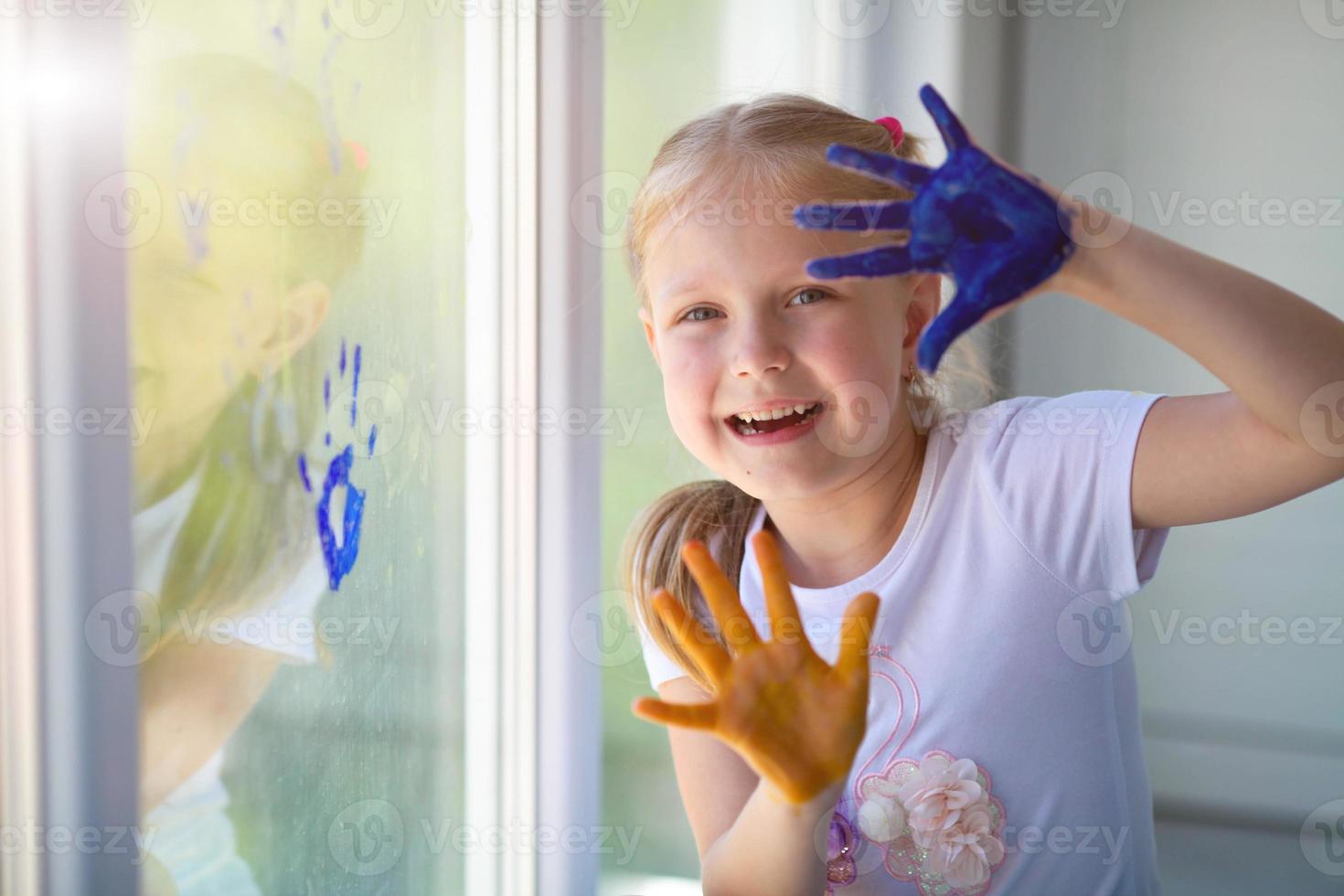 Portrait of a happy little girl draw palms on the window. Painted children's hands leave a mark on the glass. Quarantine. Stay home. photo