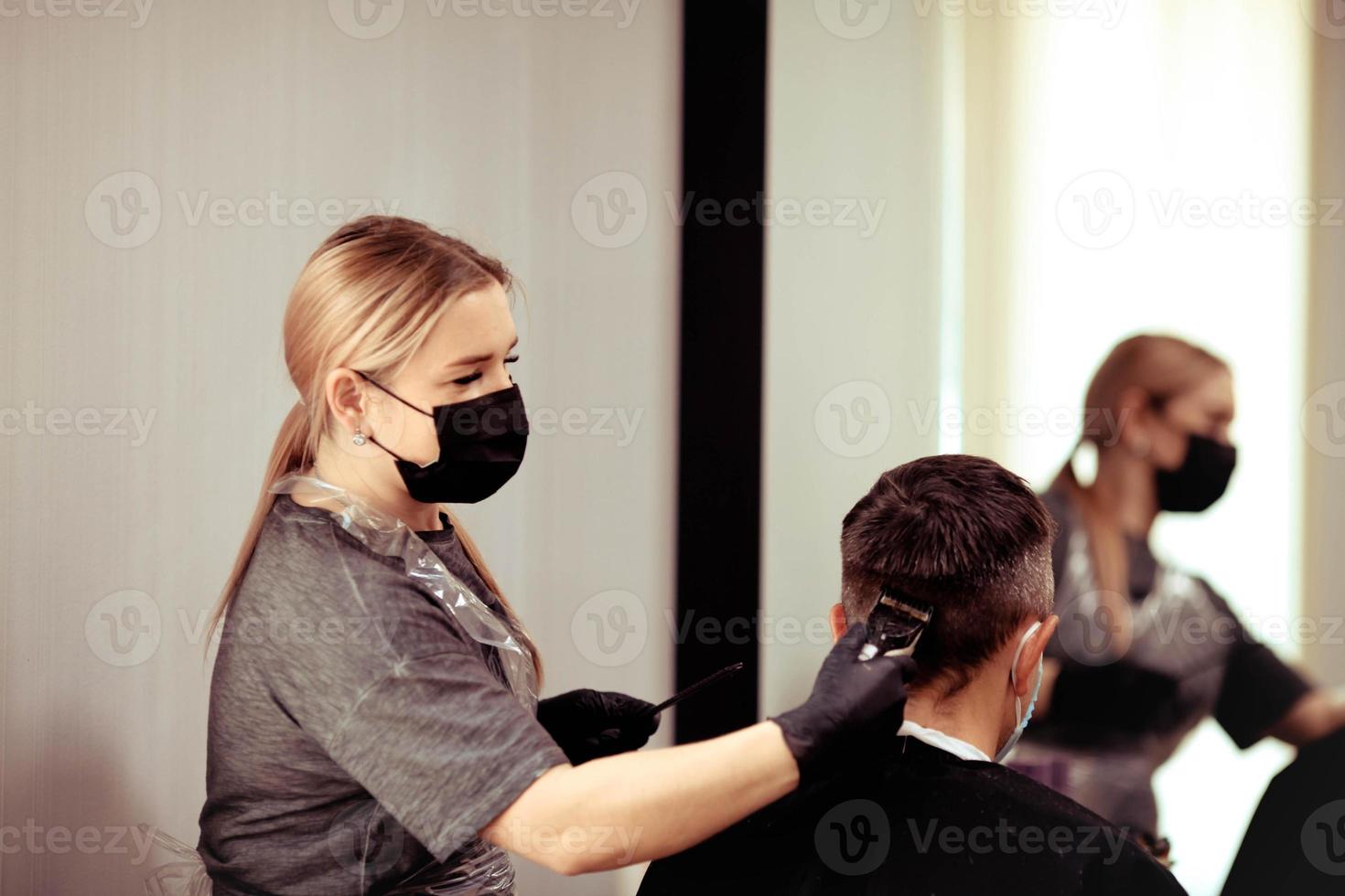 un peluquero con seguridad medidas para COVID-19, cortes un hombre en un medicina mascarilla, corte pelo con un médico máscara y caucho guantes. foto
