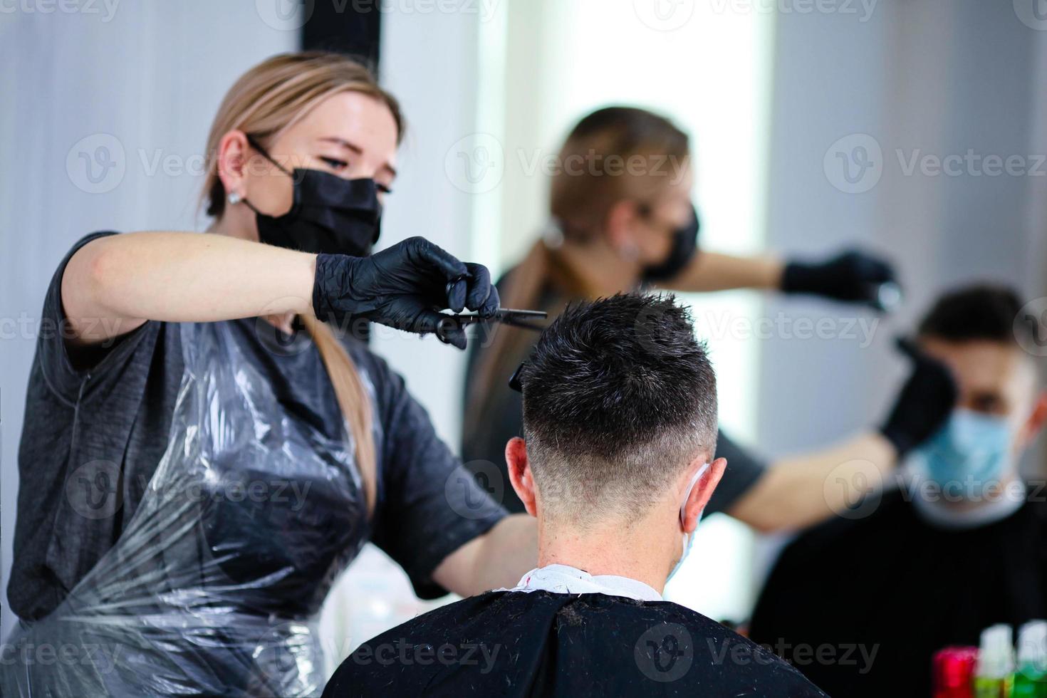 cerca arriba peluquero con seguridad medidas para COVID-19, cortes un hombre social distancia, corte pelo con un médico mascarilla. foto