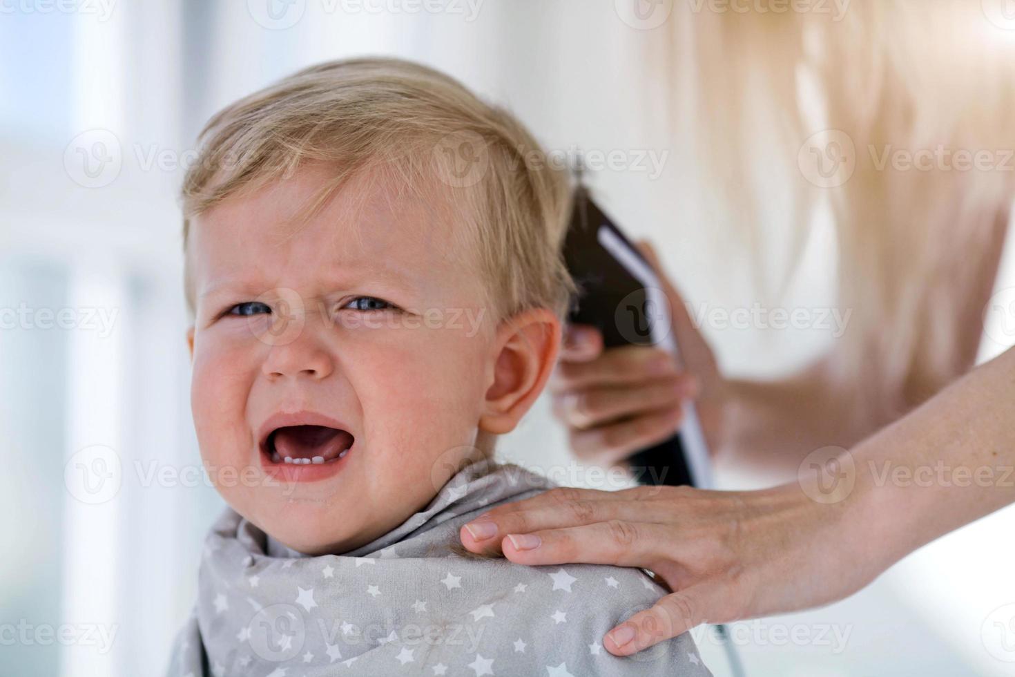 Female hands trim a crying baby with an electric hair clipper in a hairdresser. First haircut. photo