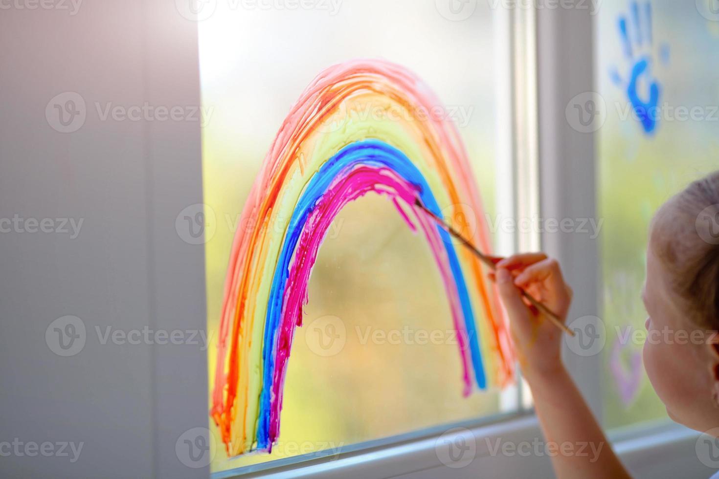 Kid painting rainbow during Covid-19 quarantine at home. Girl near window. Stay at home Social media campaign for coronavirus prevention. photo