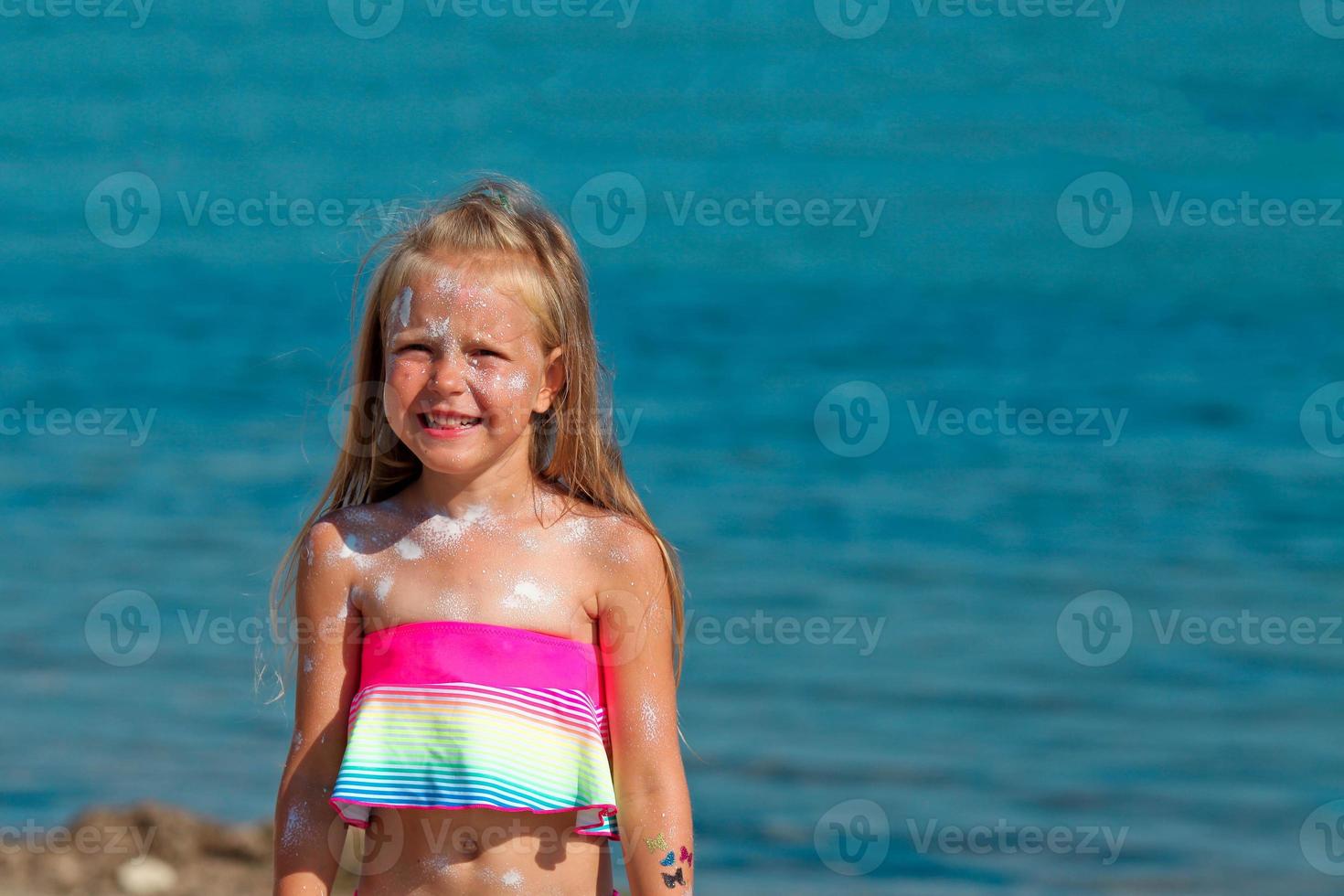 A beautiful little girl smiles in a swimsuit standing by the sea with sunscreen smeared on her face. photo