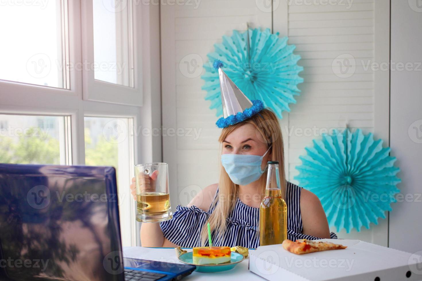 un mujer en un médico máscara se sienta en frente de un computadora portátil, celebra cumpleaños con amigos terminado el Internet, y bebidas alcohol. cumpleaños en línea foto