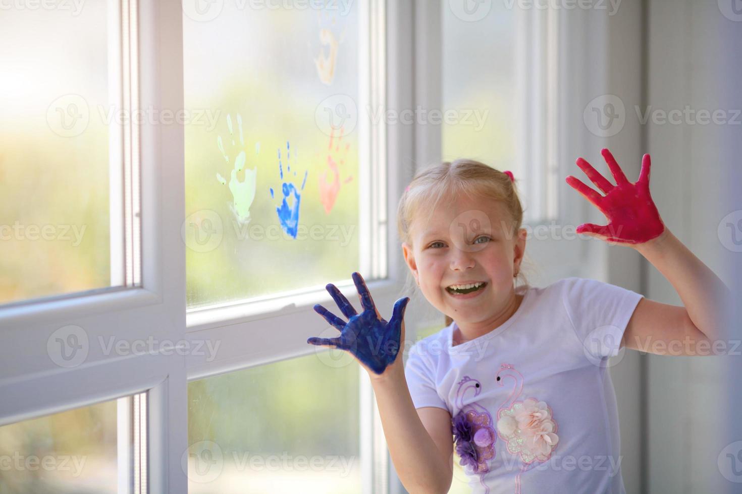 Portrait of a happy little girl draw palms on the window. Painted children's hands leave a mark on the glass. Quarantine. Stay home. photo