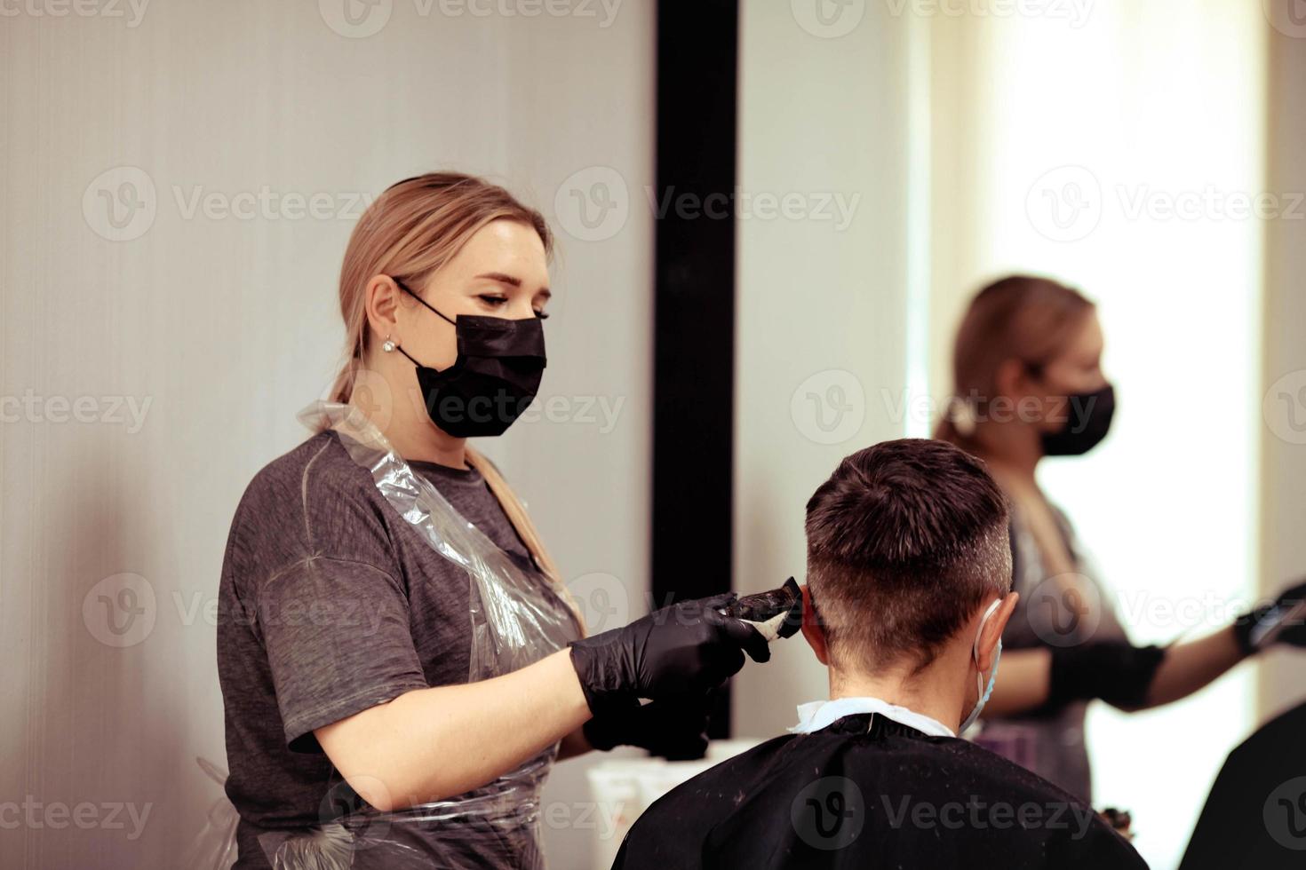 un peluquero con seguridad medidas para COVID-19, cortes un hombre en un medicina mascarilla, corte pelo con un médico máscara y caucho guantes. foto