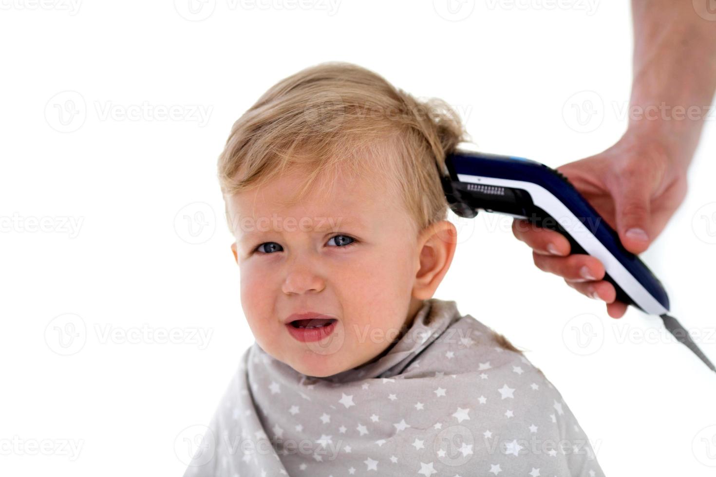 Female hands trim a crying baby with an electric hair clipper in a hairdresser. First haircut. photo