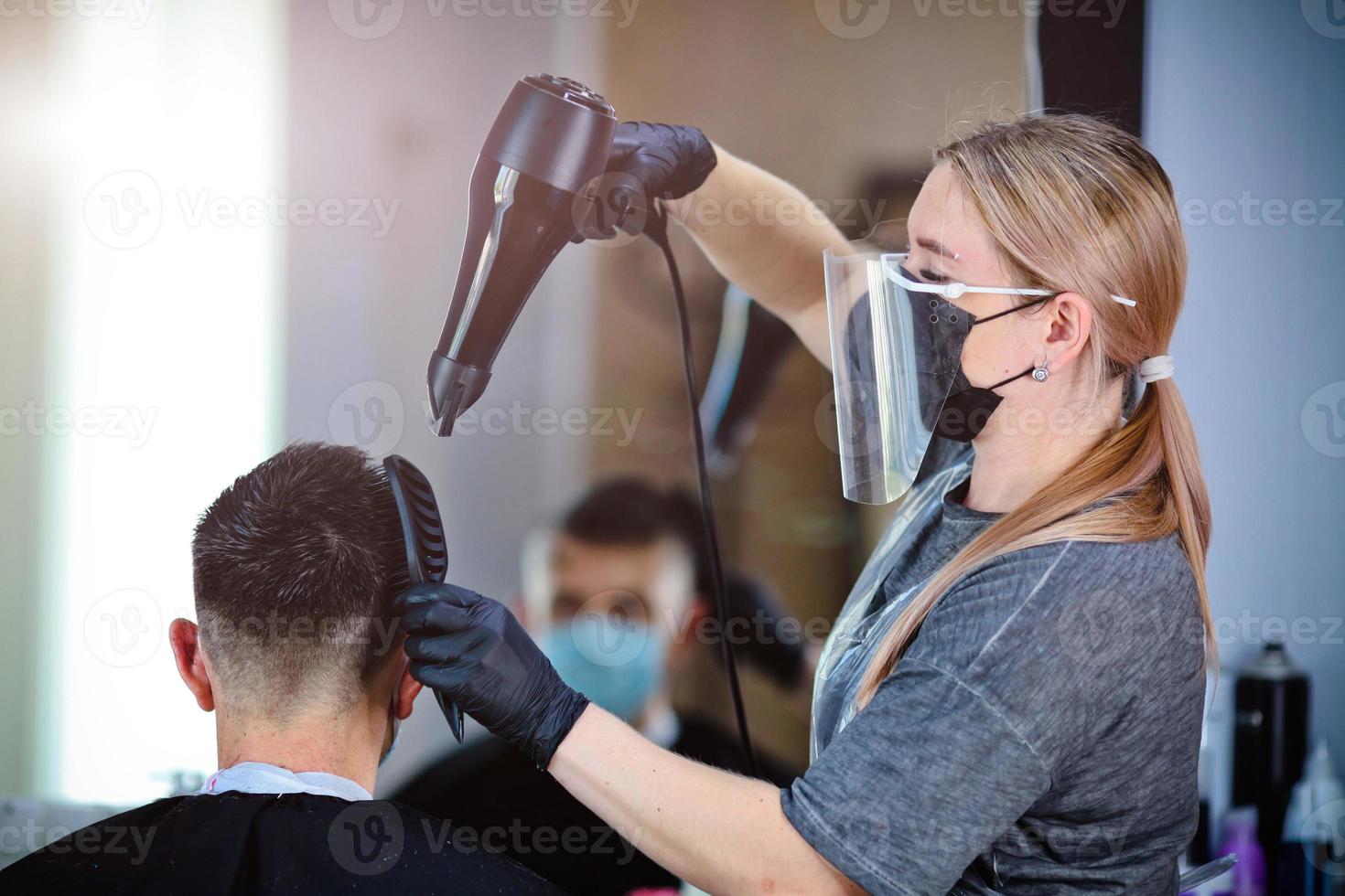 peluquero con seguridad medidas para COVID-19, golpe a secas un hombre en un medicina mascarilla, social distancia, corte pelo con un médico mascarilla, ojo máscara y caucho guantes en un belleza salón foto