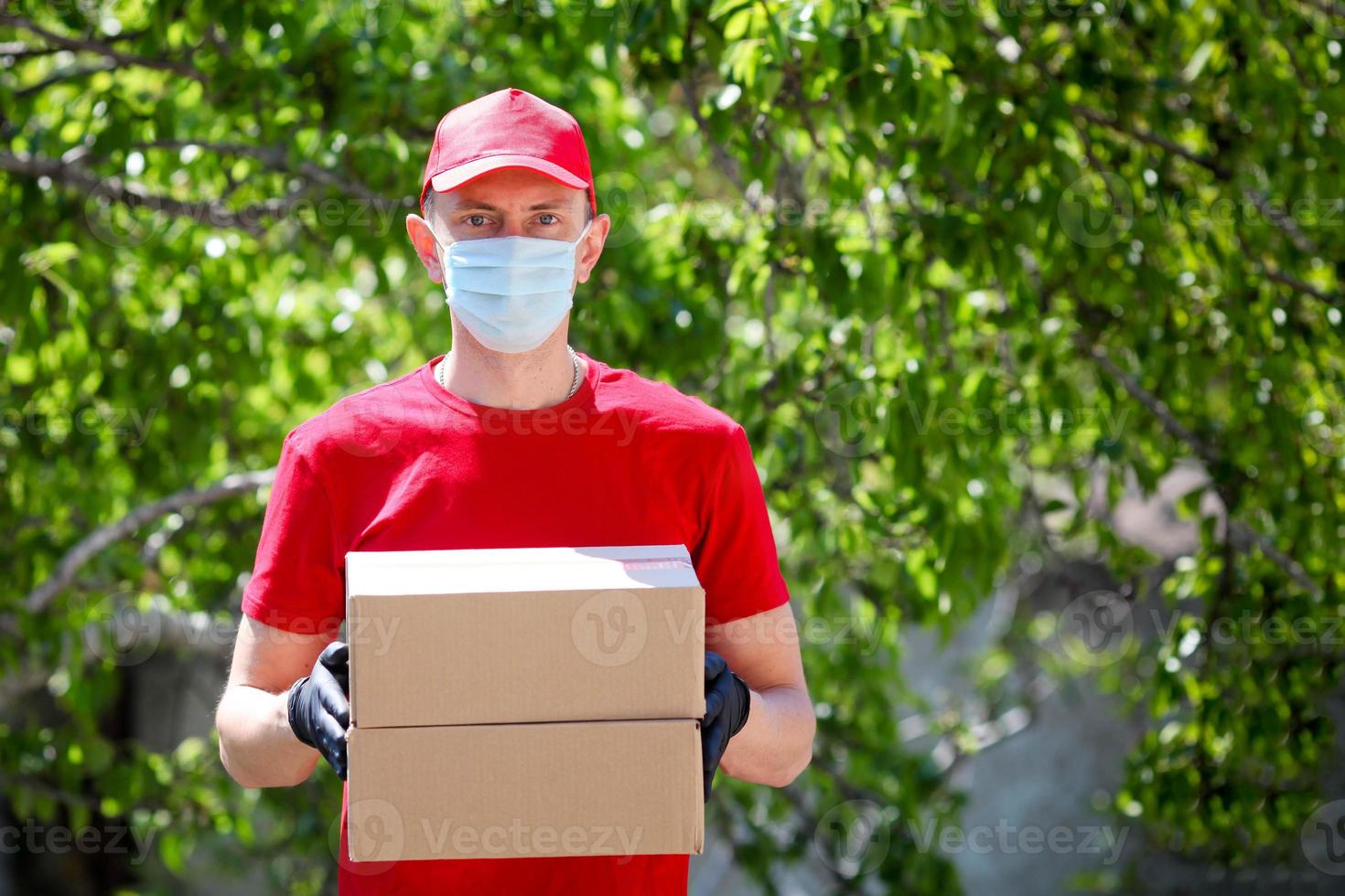 Courier in a red T-shirt and a cap in a medical mask and gloves holds a cardboard box in your hands, outdoor. photo