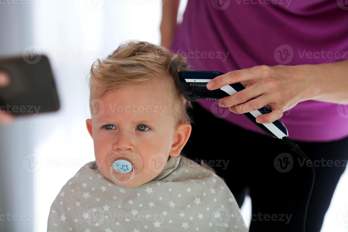 Female hands cut a child with a hair clipper in a hairdresser. A child watches cartoons on the phone and holds a dummy in his mouth. The first haircut. photo