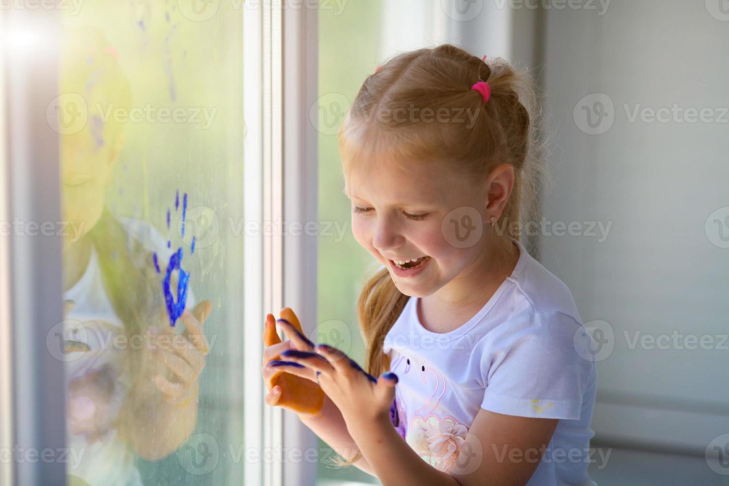 Portrait of a happy little girl draw palms on the window. Painted children's hands leave a mark on the glass. Quarantine. Stay home. photo