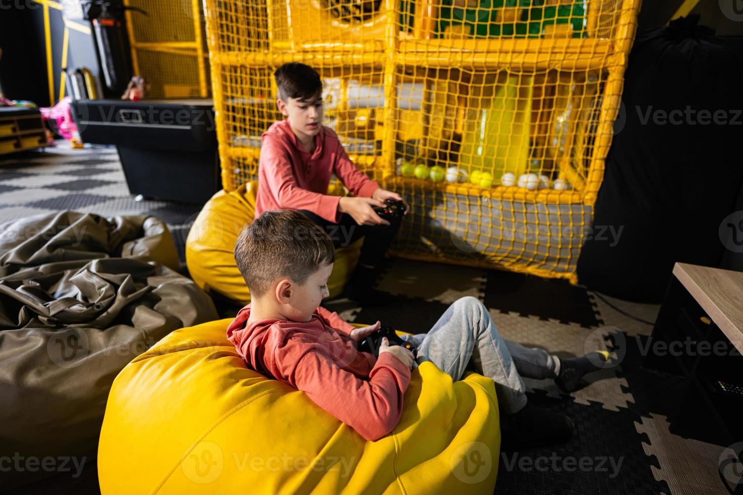 dos hermanos jugando vídeo juego consola, sentado en amarillo puff en niños jugar centro. foto