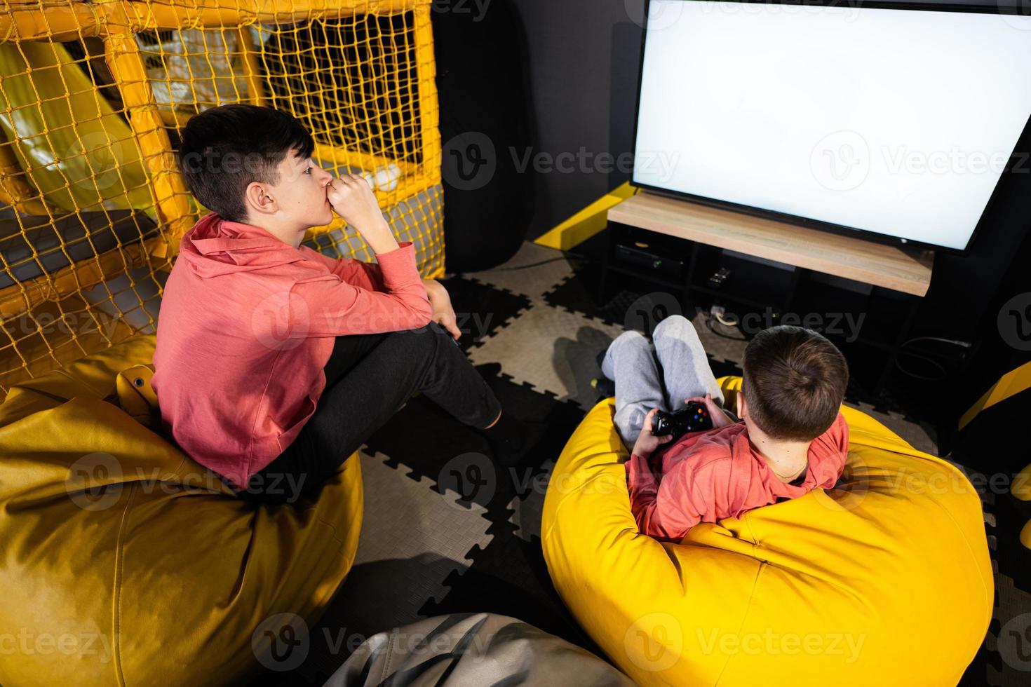 Two brothers playing video game console, sitting on yellow pouf in kids