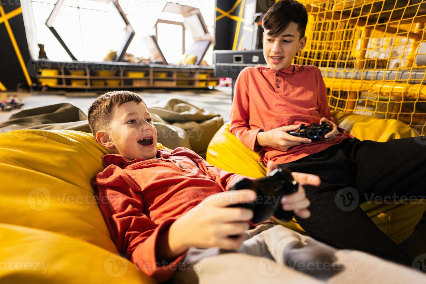 Two brothers playing video game console, sitting on yellow pouf in kids play center. photo