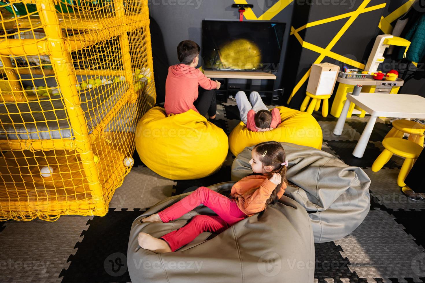Three children playing video game console, sitting on yellow pouf in kids play center. photo