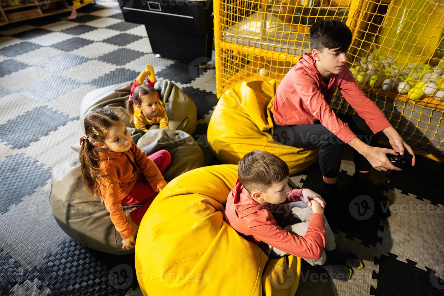 Four children playing video game console, sitting on yellow pouf in kids play center. photo