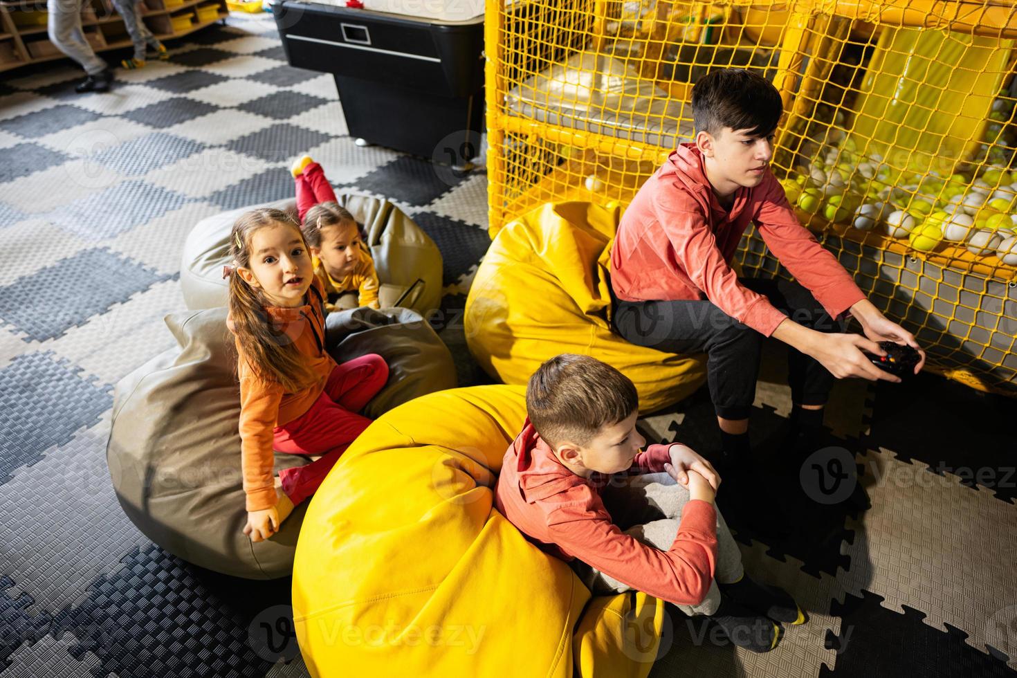 Four children playing video game console, sitting on yellow pouf in kids play center. photo