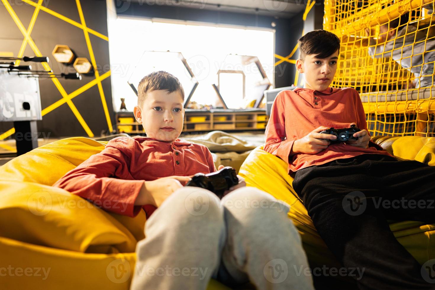 Two brothers playing video game console, sitting on yellow pouf in kids play center. photo