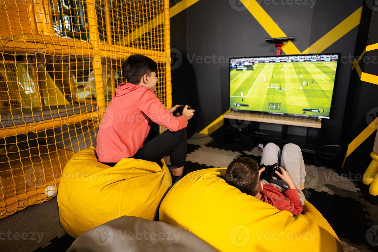 Two brothers playing football video game console, sitting on yellow pouf in kids play center. photo