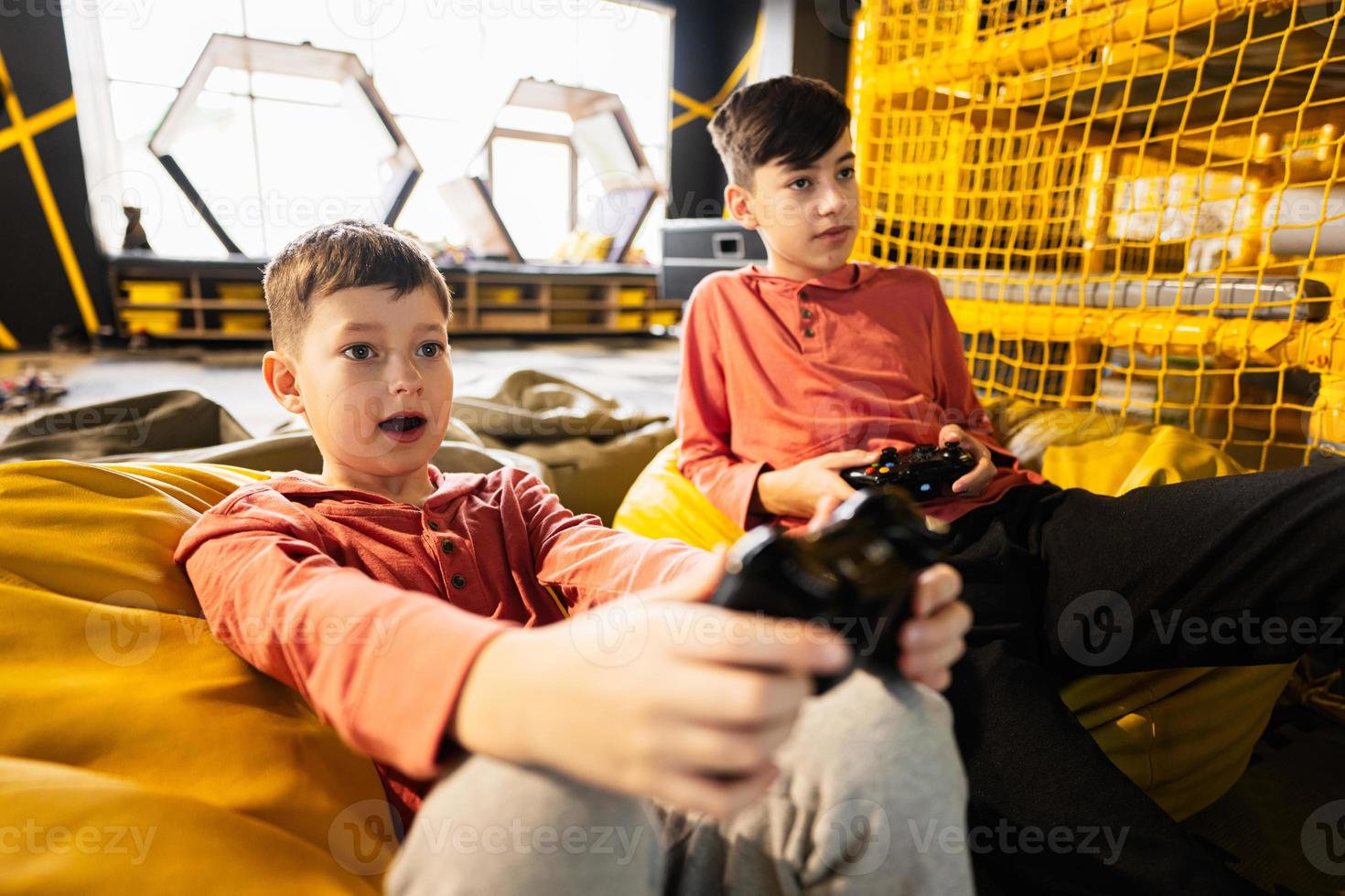 Two brothers playing video game console, sitting on yellow pouf in kids play center. photo