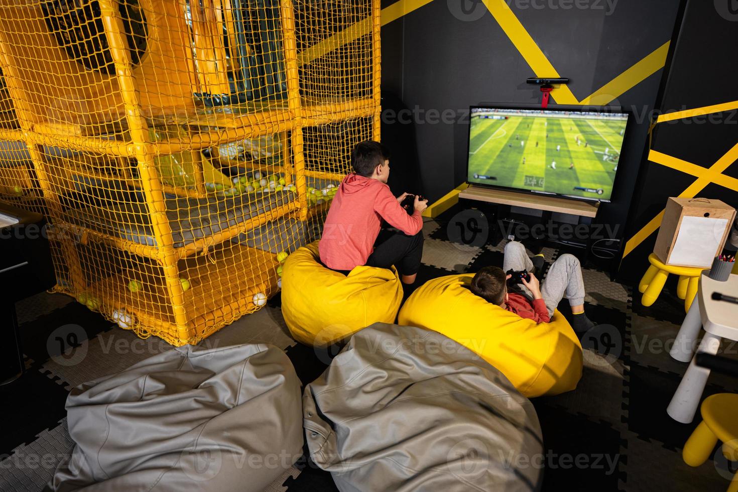 Two brothers playing football video game console, sitting on yellow pouf in kids play center. photo