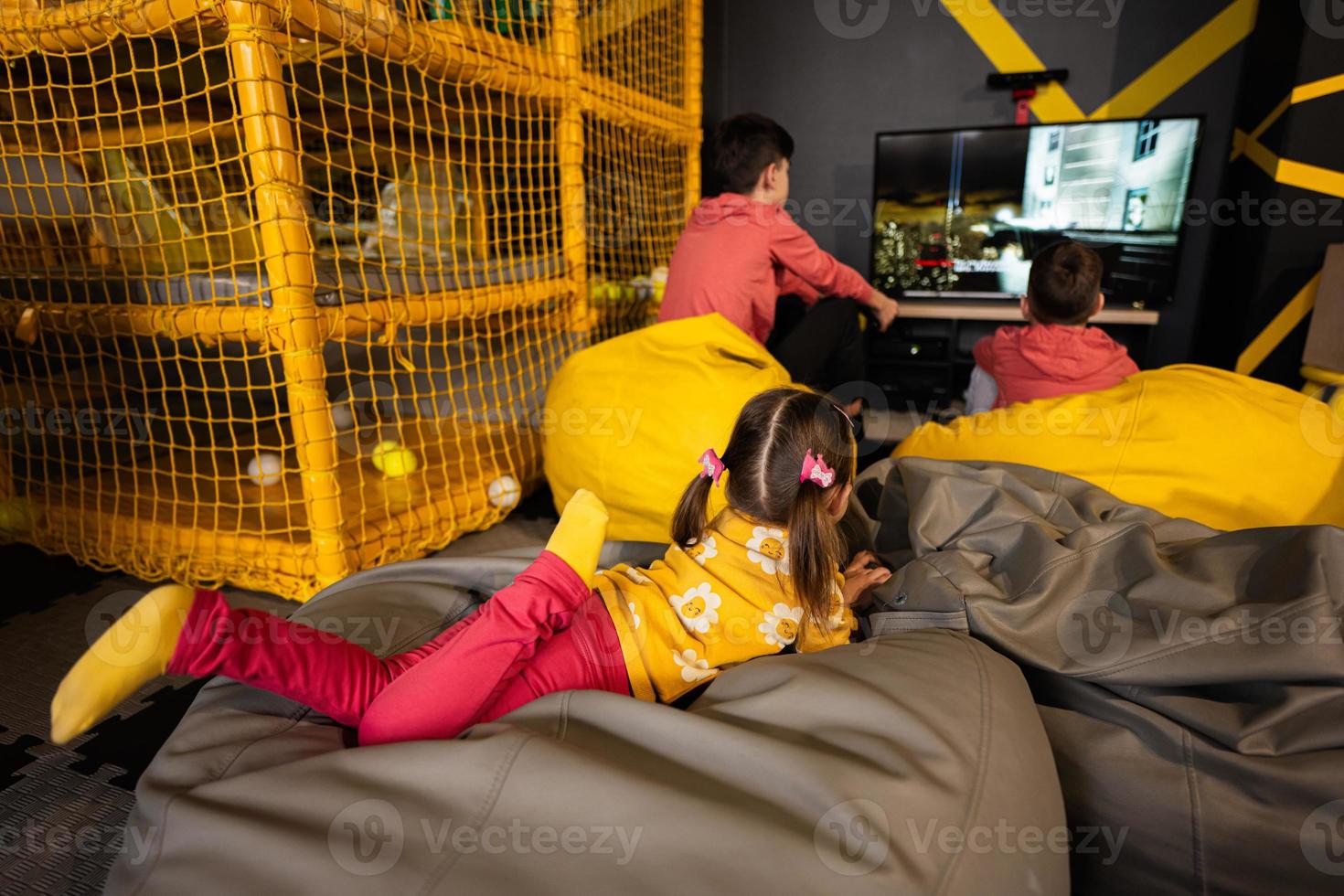 Three children playing video game console, sitting on yellow pouf in kids play center. photo