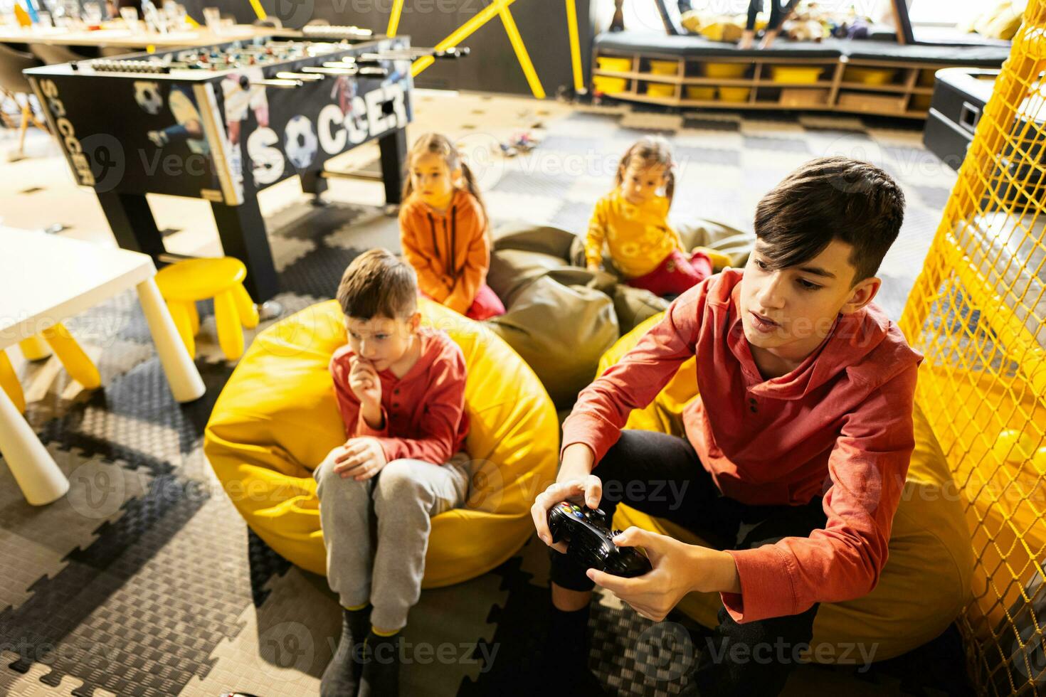 Four children playing video game console, sitting on yellow pouf in kids play center. photo