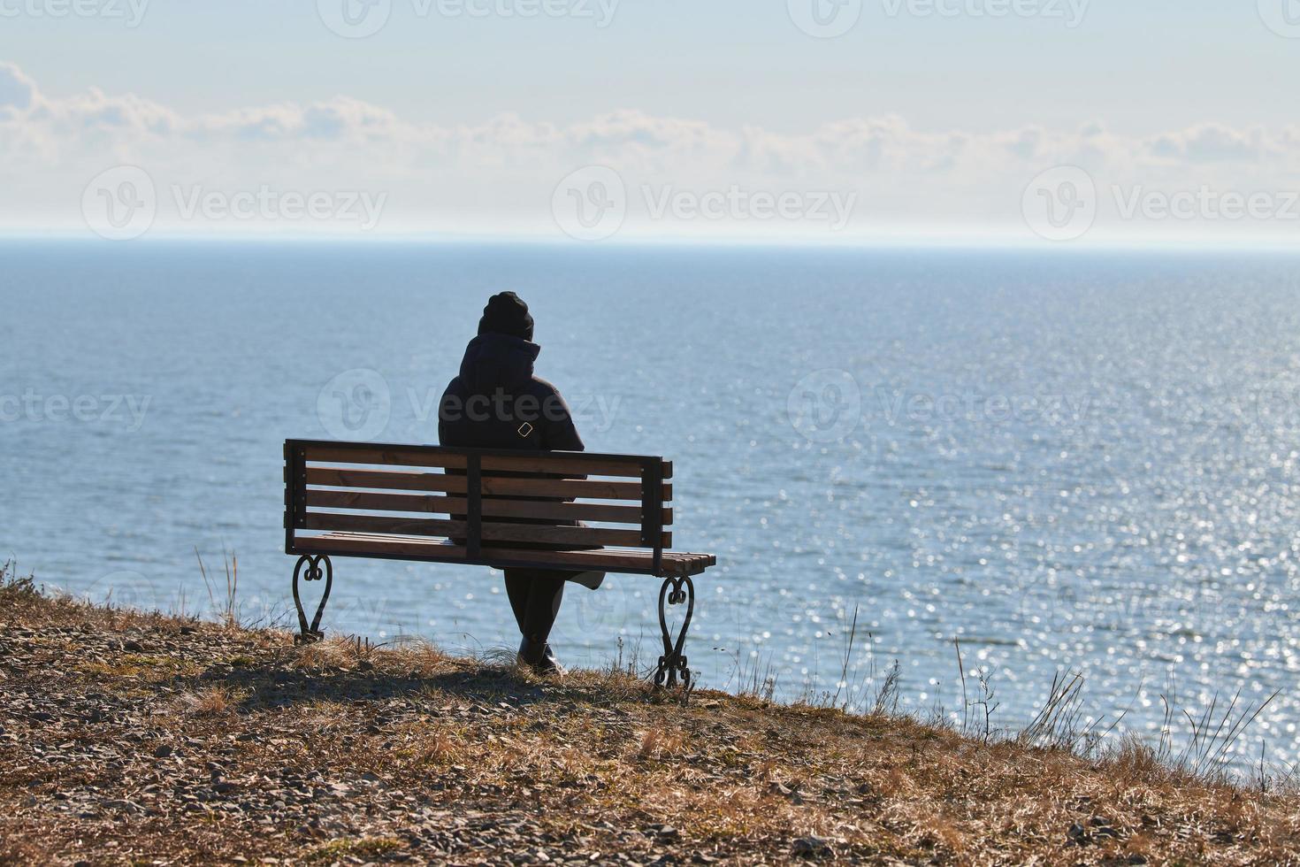 una chica soltera con una chaqueta negra y un sombrero sentada en un banco en un acantilado frente al mar, un lugar tranquilo y tranquilo foto